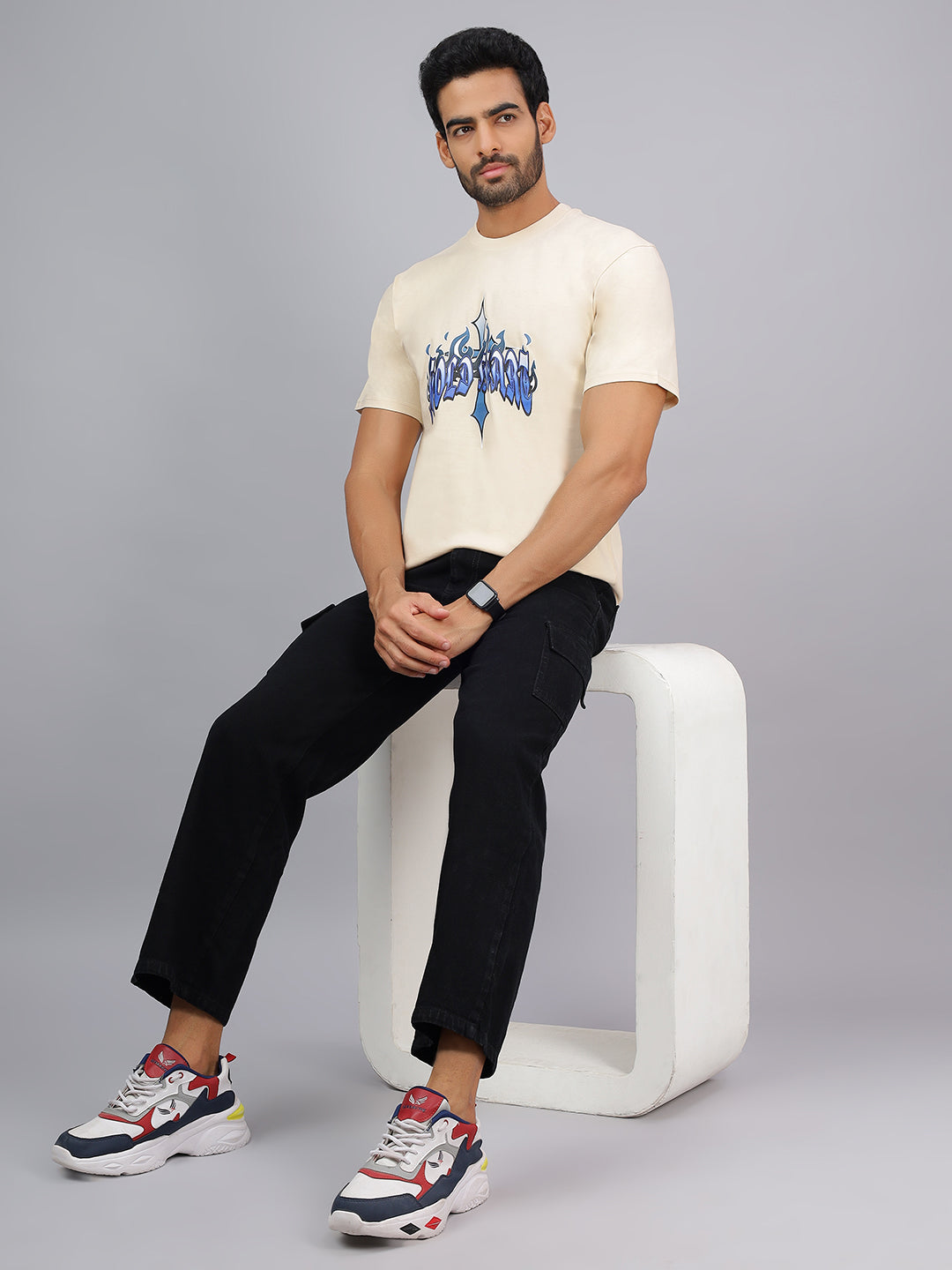 Man sitting on a white stool wearing a cream t-shirt with blue graphics, black pants, and colorful sneakers on a gray background.