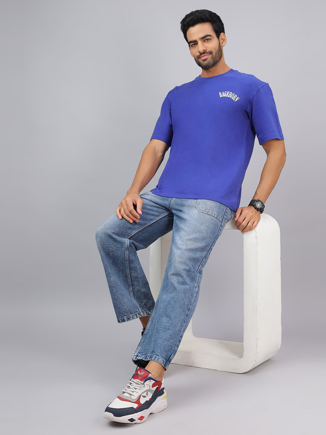 Man wearing a blue t-shirt and jeans sitting on a white stool against a gray background