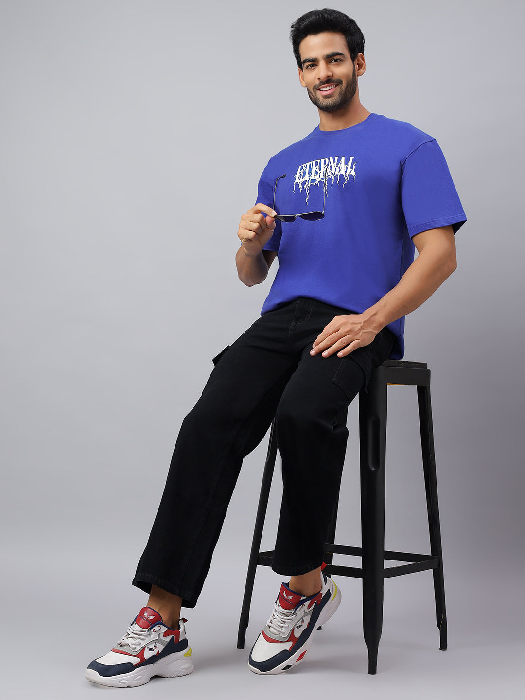 Man sitting on a stool wearing a blue t-shirt with text and black pants, against a gray background